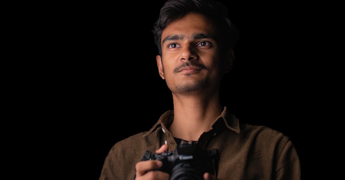 Portrait of a young man holding a DSLR camera in a dark studio setting.