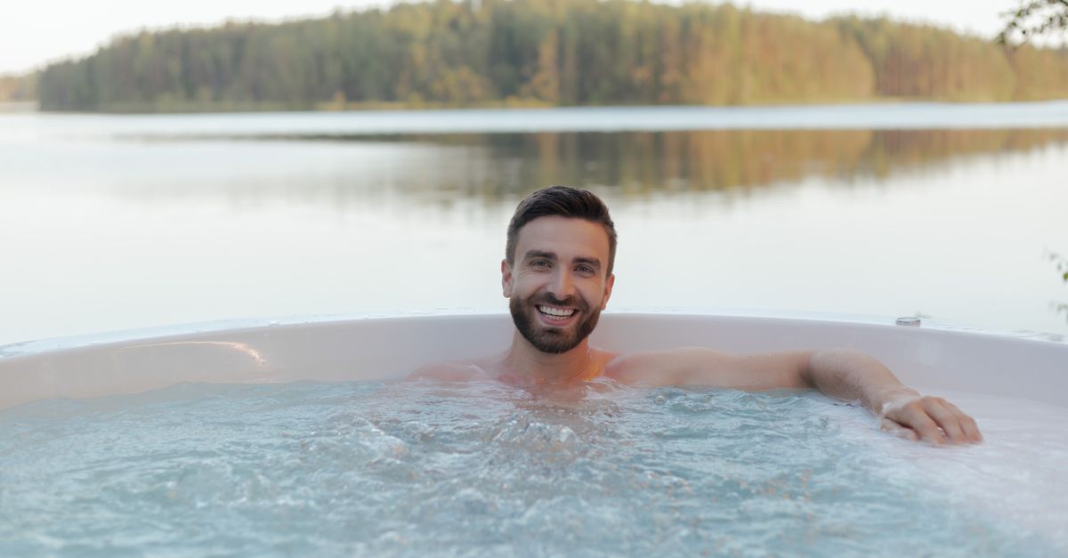 Shirtless man smiling in an outdoor hot tub by a serene lake, enjoying relaxation and wellness.