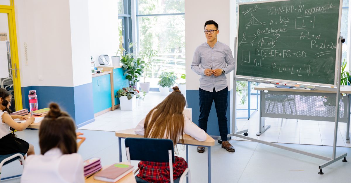 Teacher instructing students in a bright classroom with a blackboard filled with math equations.