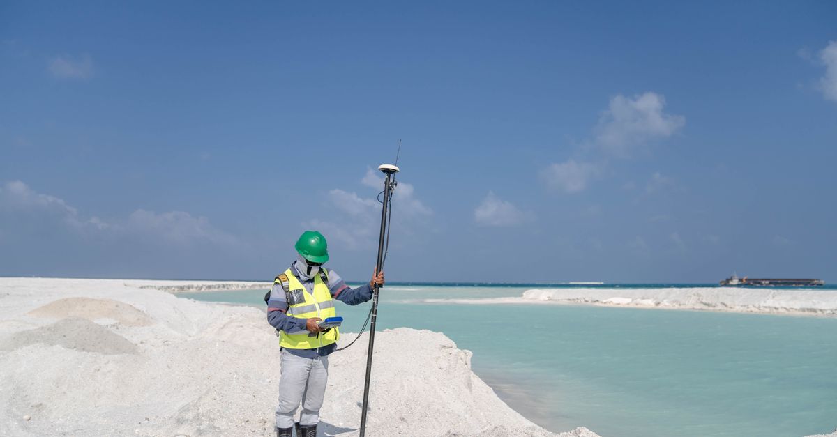Surveyor in safety gear measuring near a turquoise lake under a clear sky.