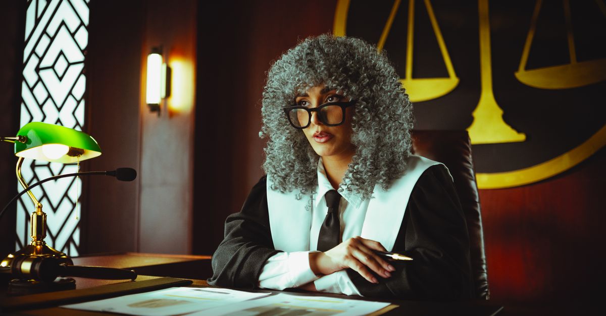 Female judge in courtroom setting, sitting at desk with justice scales in background.