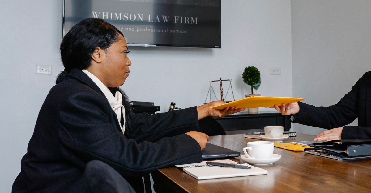 Side view of African American woman in formal wear sitting at table with documents and laptop while surrendering documents to colleague