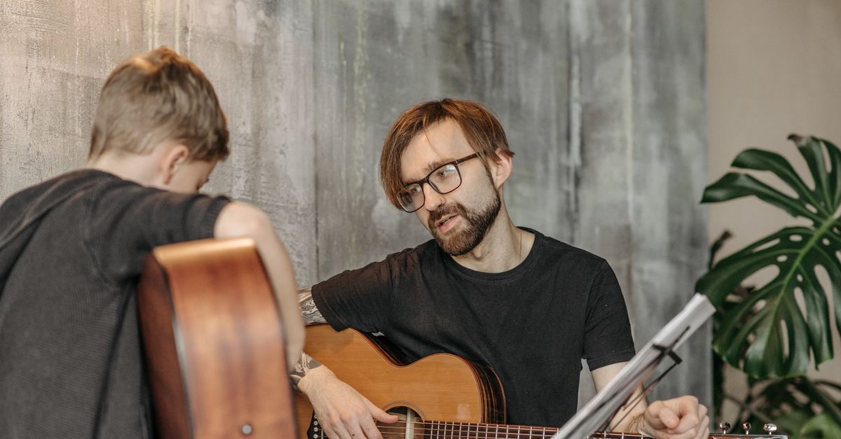 A young boy learning guitar from a bearded man indoors, emphasizing musical guidance.