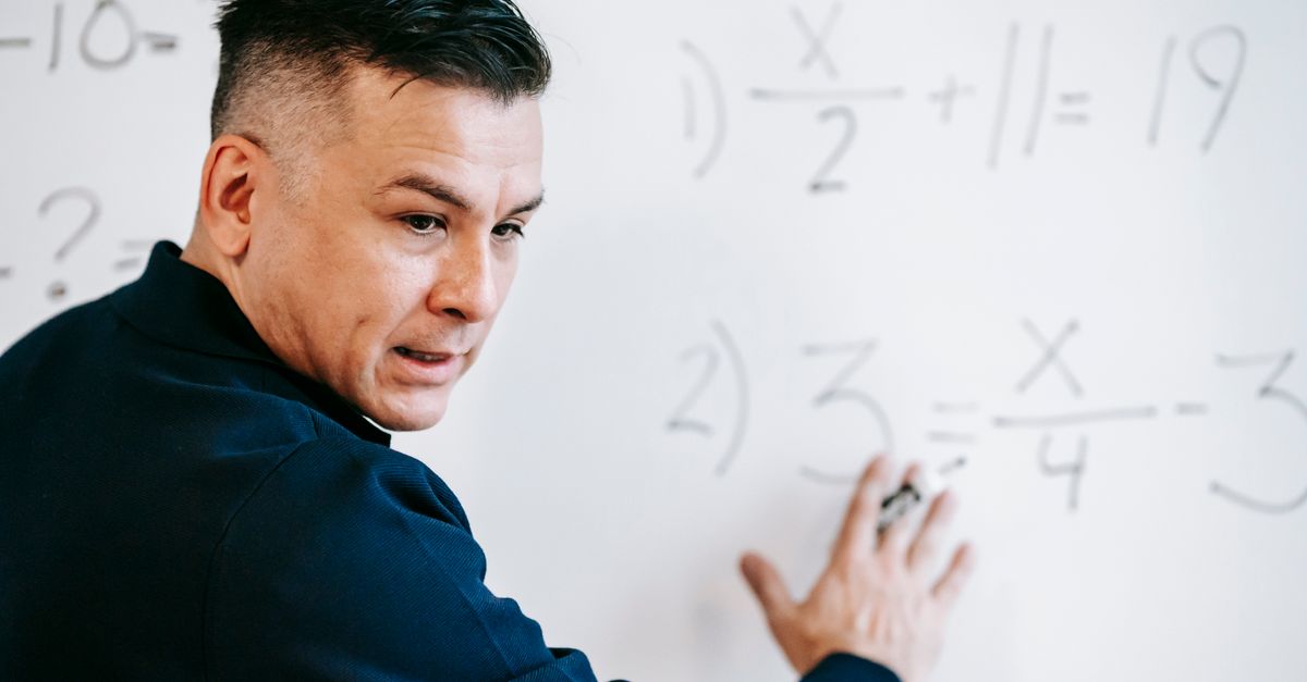 A mathematics instructor explaining algebraic equations on a whiteboard in a classroom setting.