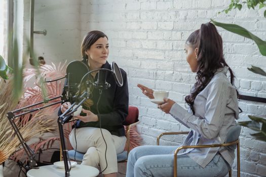 Serious host listening to African American female guest giving interview while sitting with cup of coffee near mic