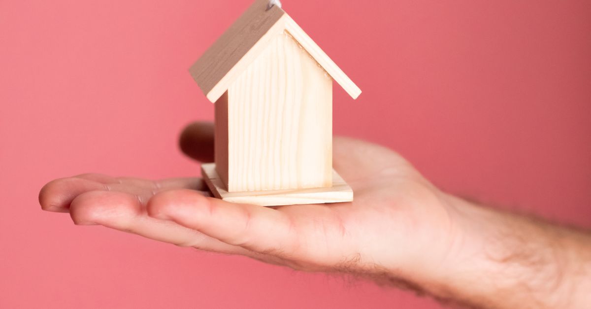 Wooden house model in hand symbolizing real estate concepts on pink background.