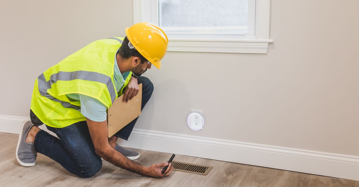 A construction worker in a hard hat inspects a floor vent indoors, ensuring quality and safety standards.
