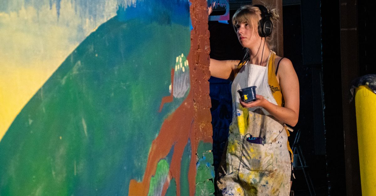 Female artist painting a vibrant mural while listening to music in an indoor studio environment.