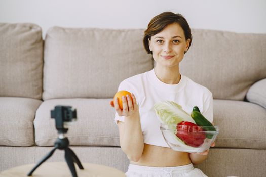Optimistic female in white clothes sitting near sofa and holding bowl with fresh vegetables and orange in hands and looking at camera
