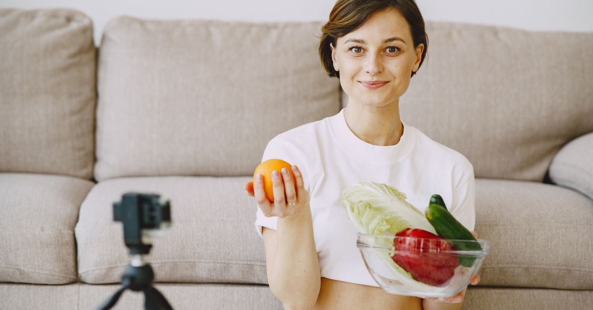 Optimistic female in white clothes sitting near sofa and holding bowl with fresh vegetables and orange in hands and looking at camera