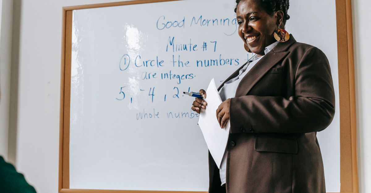 Crop smiling African American female teacher standing near whiteboard with papers in hands during math lesson