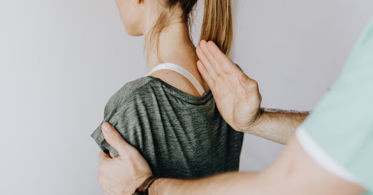Back view of crop unrecognizable osteopath in uniform and wristwatch checking up back of slim female patient in casual wear on white background