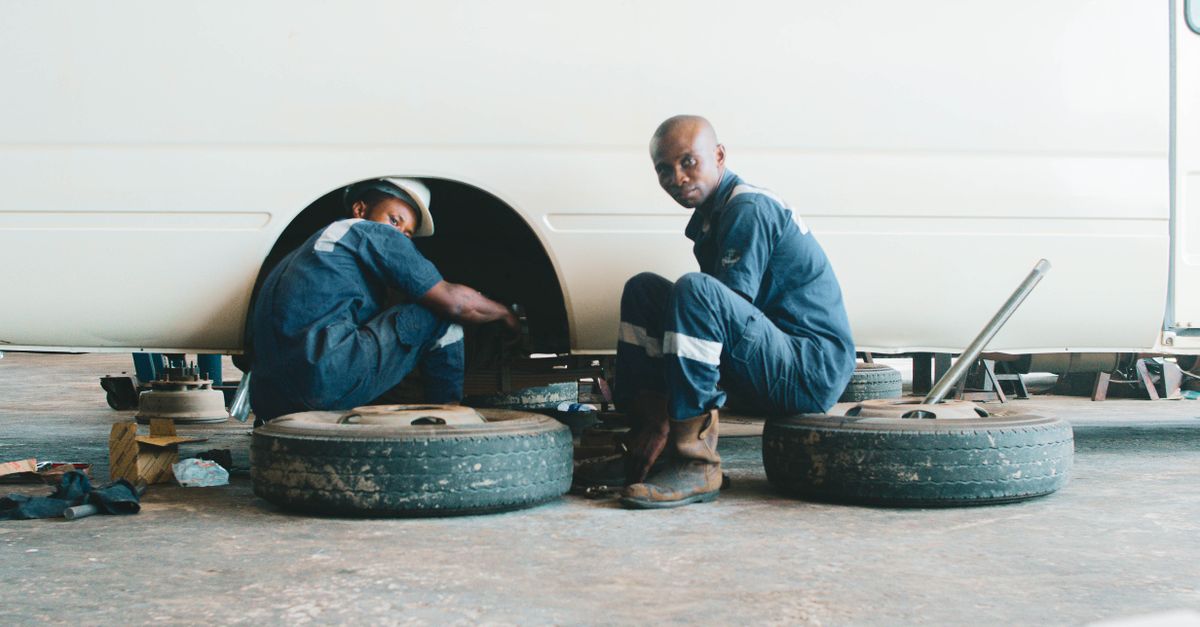 Two mechanics in coveralls repairing vehicle in Nigerian workshop, focusing on wheel maintenance.