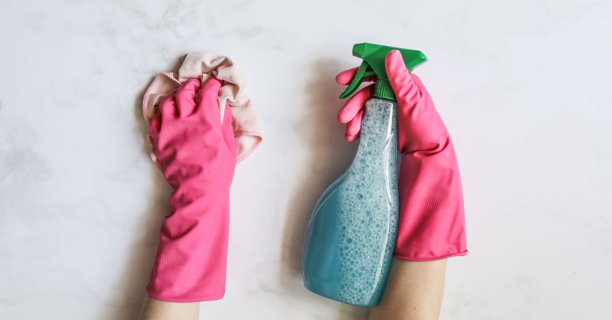 A pair of hands in pink gloves are cleaning with a spray bottle on a white background.