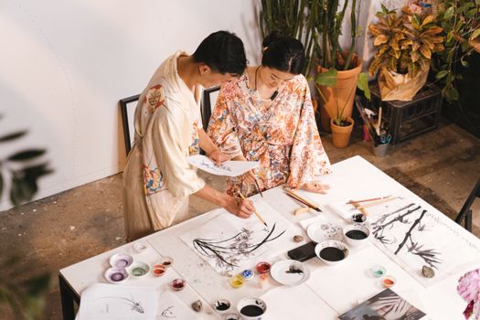 Two Asian artists in robes collaborating on a painting in an art studio using various brushes and paints.