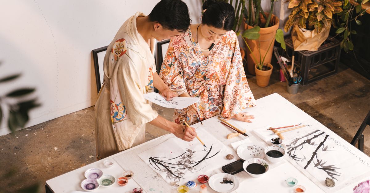 Two Asian artists in robes collaborating on a painting in an art studio using various brushes and paints.