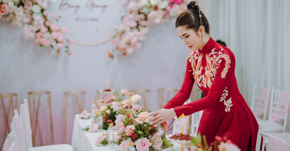 An Asian woman in traditional red dress arranges a floral centerpiece at an elegant indoor event.