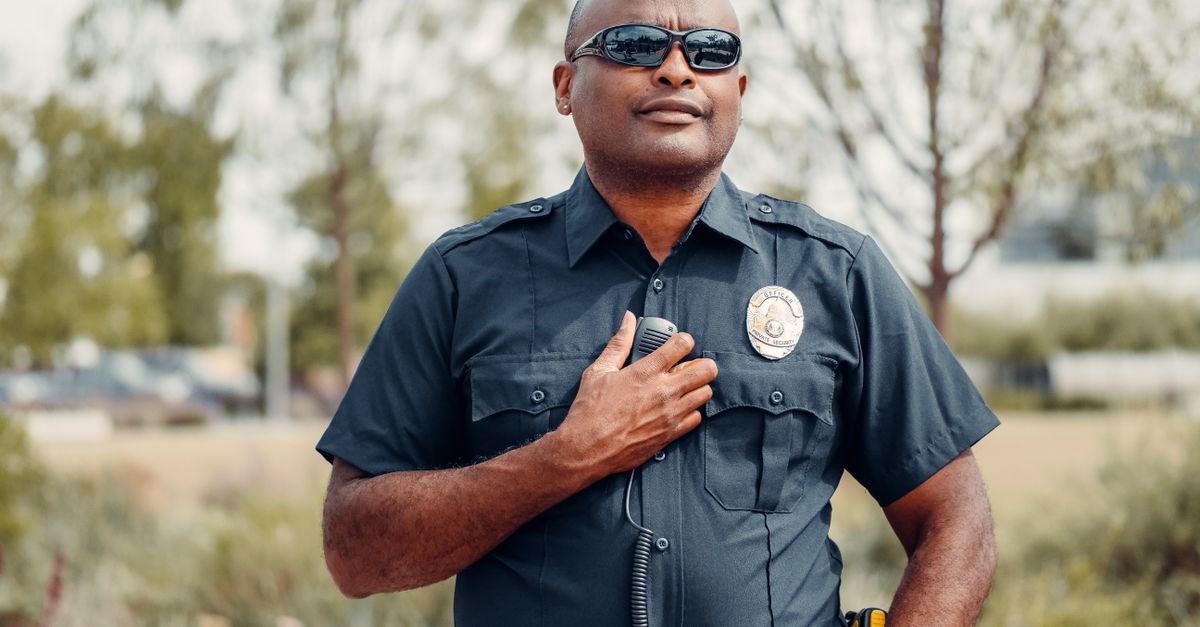 Confident police officer in uniform outdoors, hand on badge, sunny park setting.