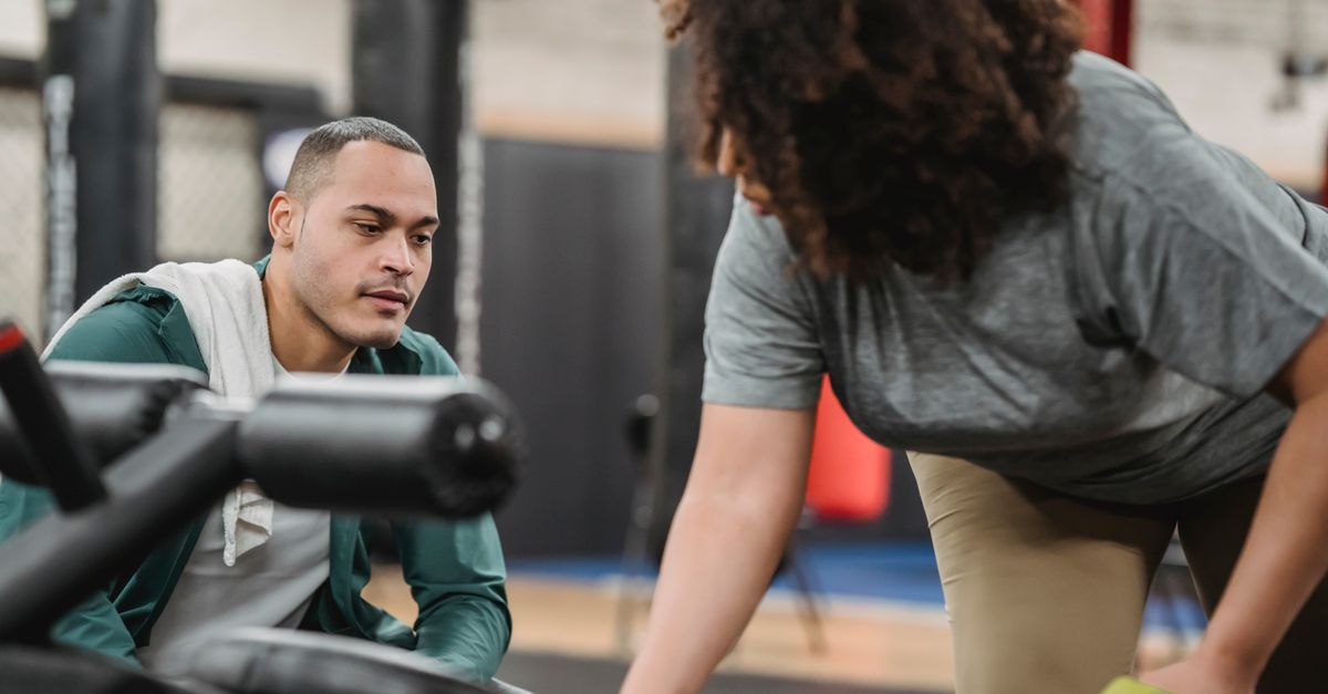 Experienced instructor watching anonymous African American female lifting dumbbell on blurred background of gym