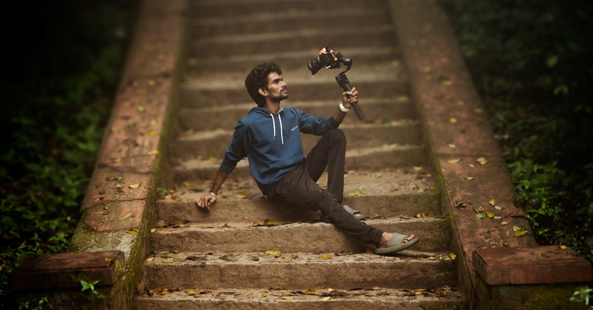 A man in a blue hoodie posing on outdoor steps while holding a camera on a gimbal, surrounded by greenery.