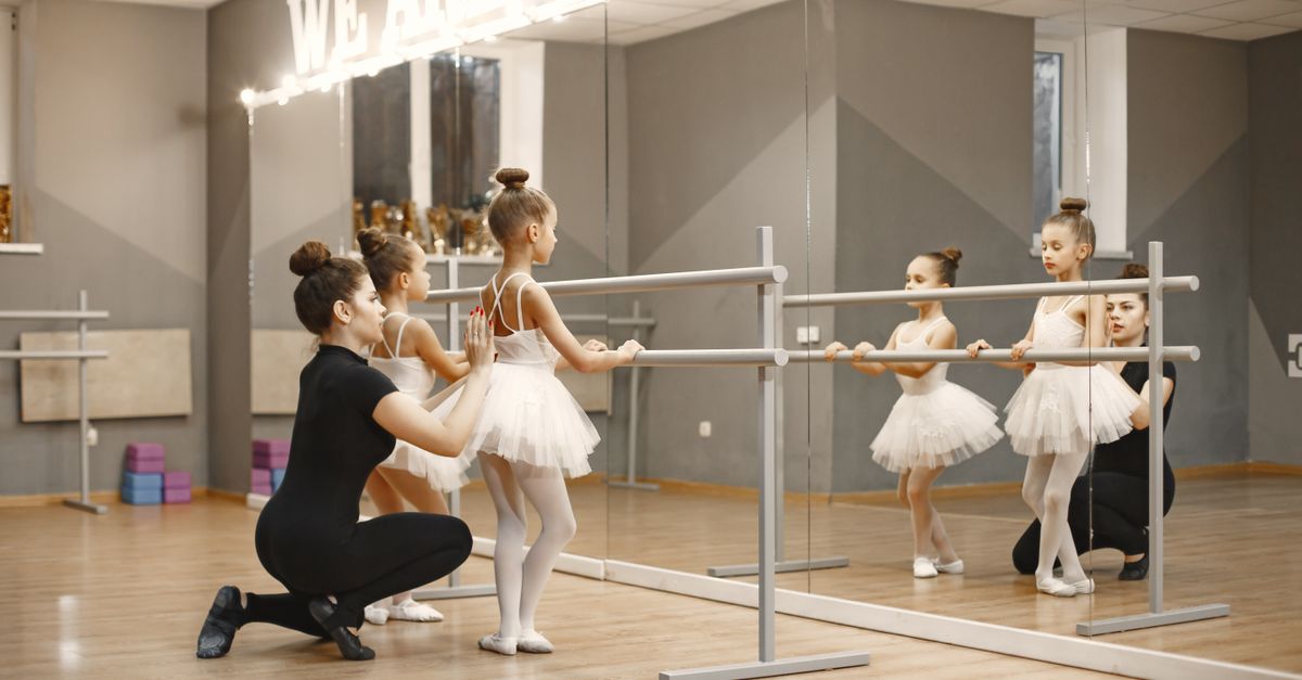 Ballet instructor helps young girls in tutus practice at a mirrored barre.