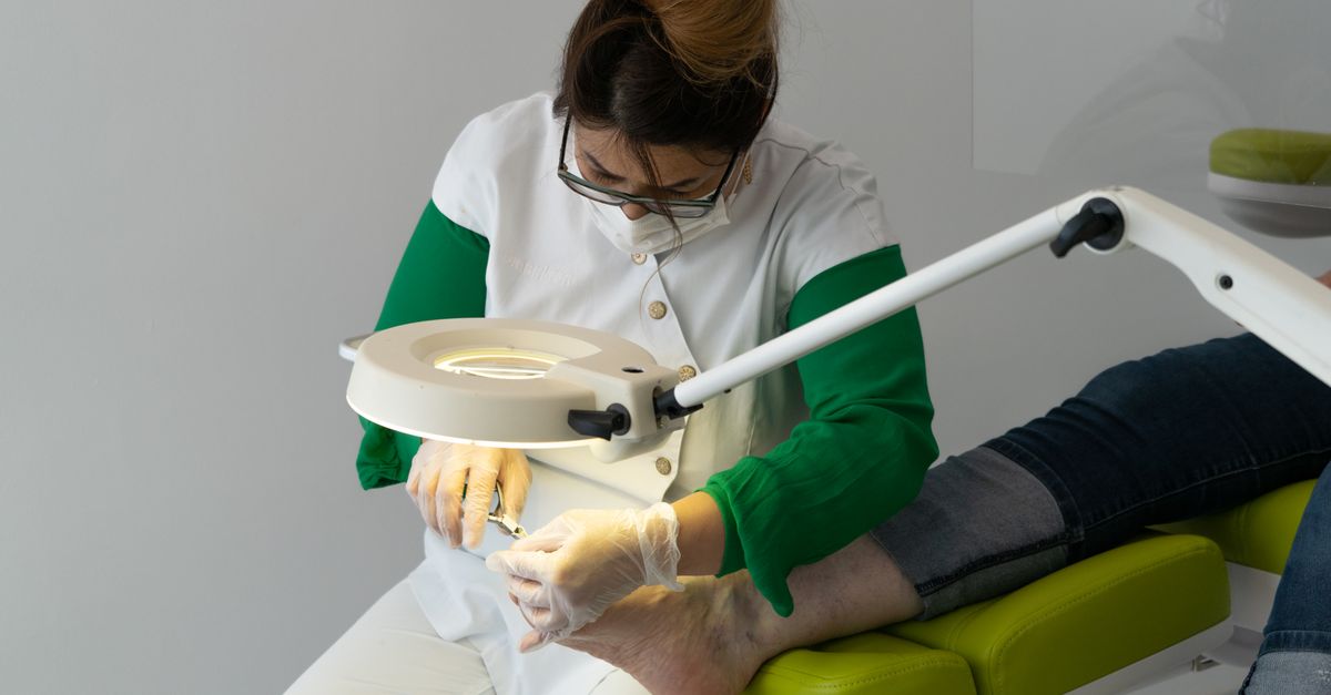 Concentrated female master in protective mask and gloves making pedicure for client sitting on couch