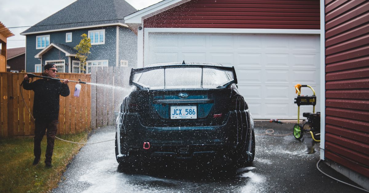 Full body of young male in black wear using long thin hosepipe while washing luxury vehicle in yard of tall private house