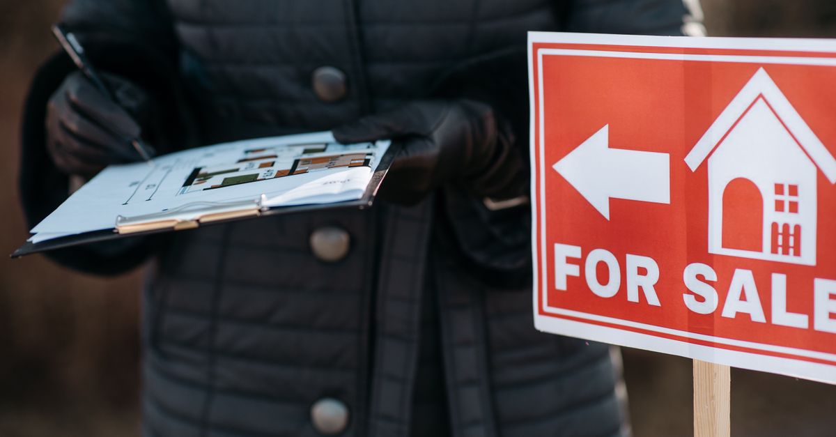 A real estate agent reviews plans next to a 'For Sale' sign outdoors, indicating property availability.