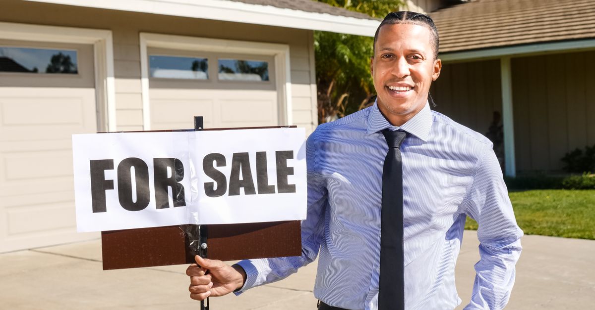 Smiling real estate agent with a for sale sign in front of a house.