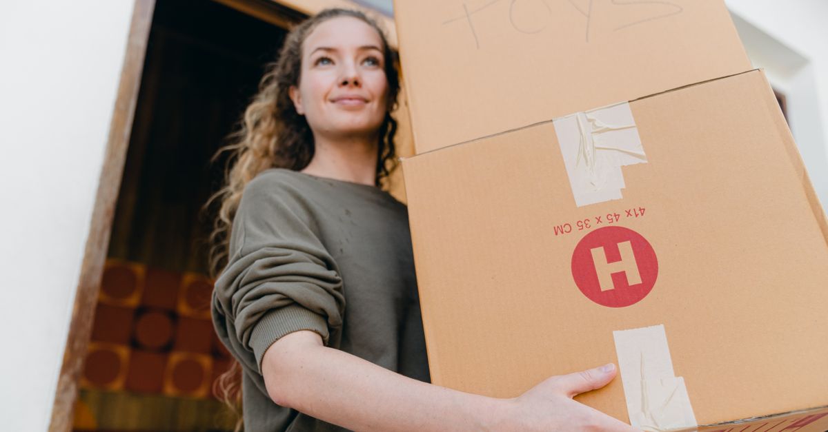 Confident young lady in casual wear carrying heavy carton boxes with toys and looking away while leaving old house