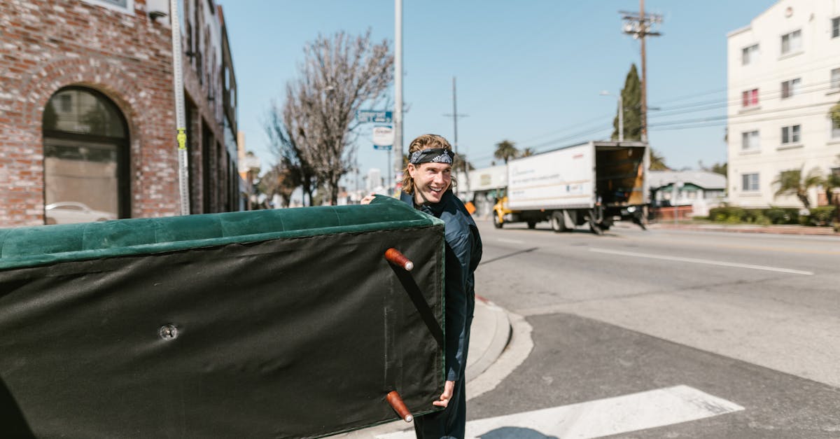 A cheerful man carries a large couch across a sunny urban street during a move.