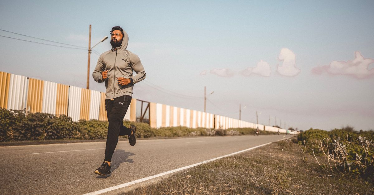 A bearded man in a hoodie jogs along an empty road under a clear sky.