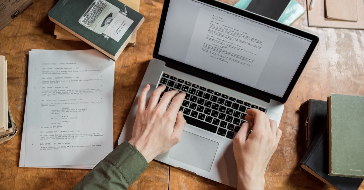 High-angle view of hands typing on a laptop surrounded by books and papers.
