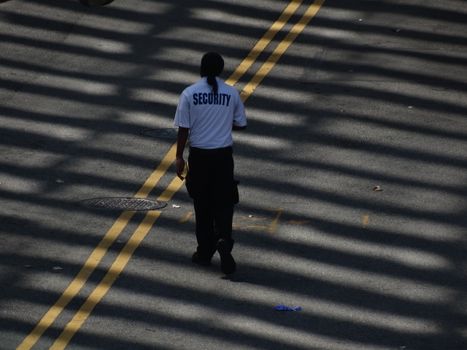 A security guard walks down a sunlit street, casting shadows on the pavement.