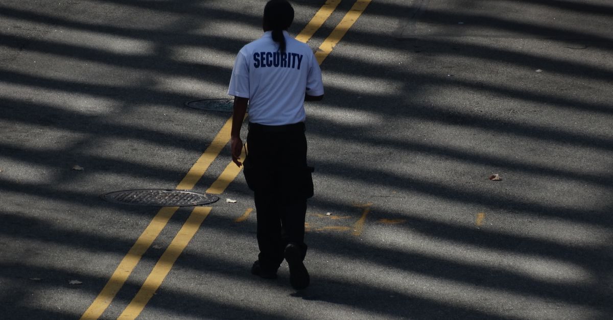 A security guard walks down a sunlit street, casting shadows on the pavement.