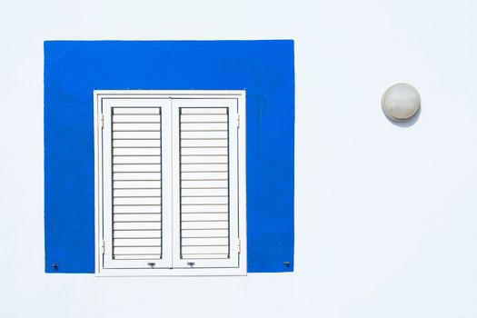A minimalist facade featuring a white wall with a blue-framed shuttered window and a round lamp.