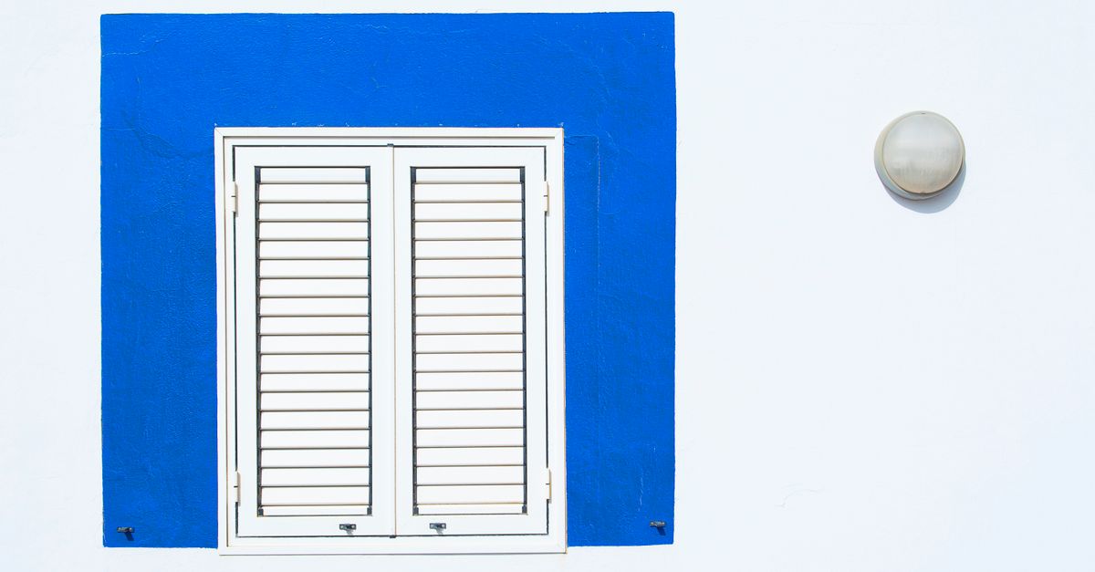 A minimalist facade featuring a white wall with a blue-framed shuttered window and a round lamp.