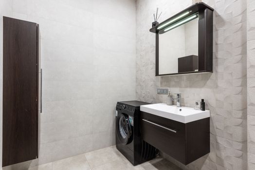 Modern black washing machine placed at wall near sink with dispenser and mirror in stylish light bathroom with brown cupboard