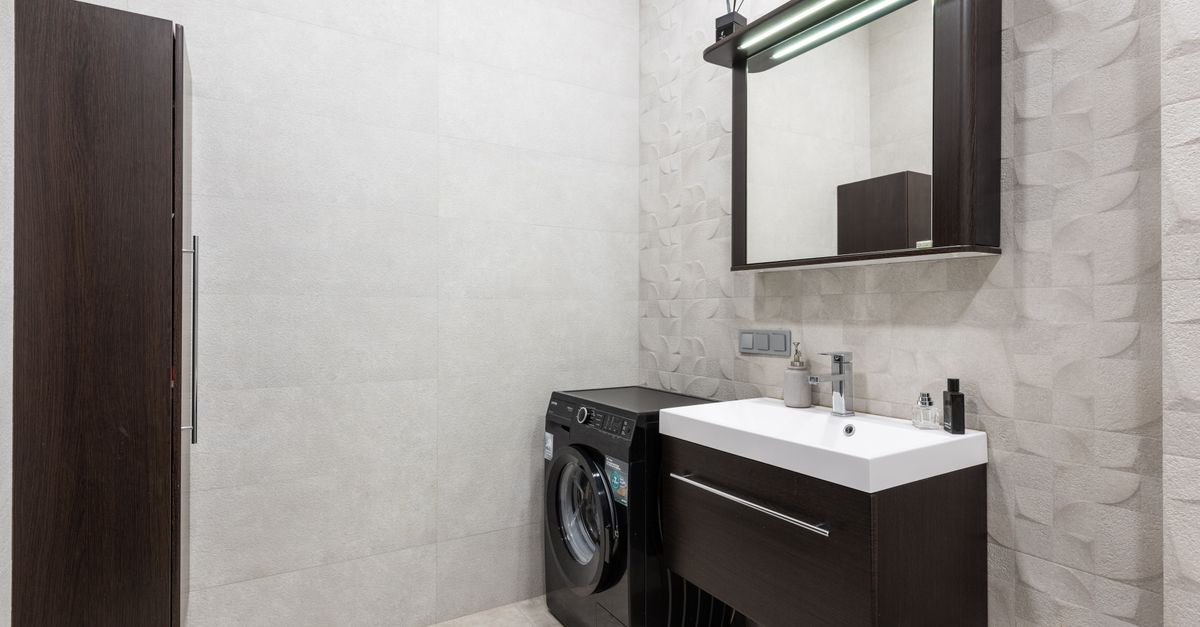 Modern black washing machine placed at wall near sink with dispenser and mirror in stylish light bathroom with brown cupboard