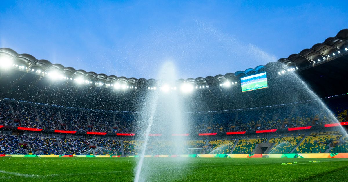 Nighttime view of a soccer stadium with sprinklers watering the field, enhancing the vibrant atmosphere.
