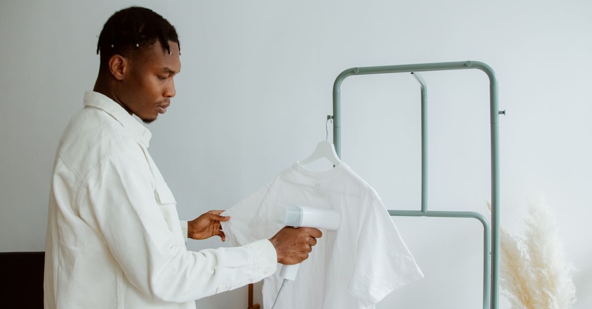 A man uses a steamer to freshen a white shirt on a clothing rack in a minimalist home interior.