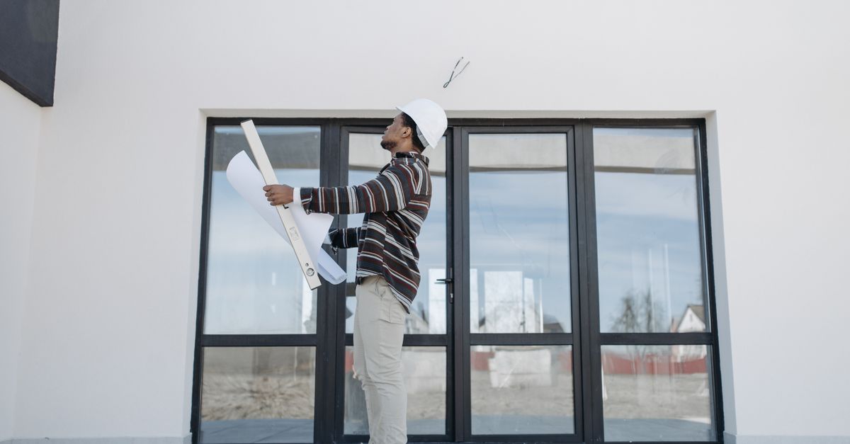 A black architect wearing a white helmet reviews construction plans outside a modern building.