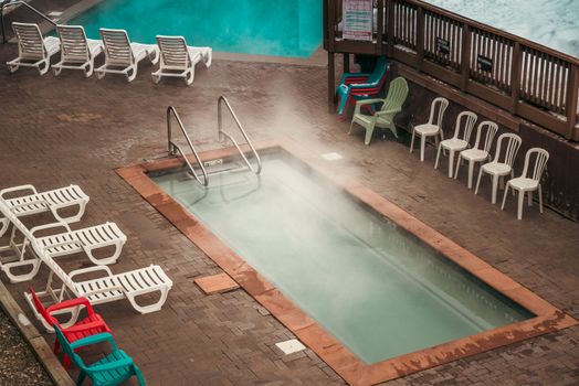 Aerial view of a steaming swimming pool surrounded by sunbeds and colorful chairs on a paved area.