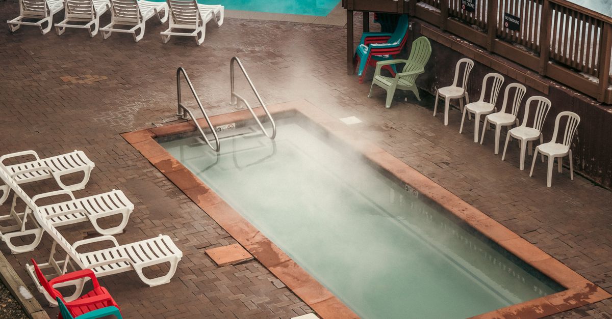 Aerial view of a steaming swimming pool surrounded by sunbeds and colorful chairs on a paved area.