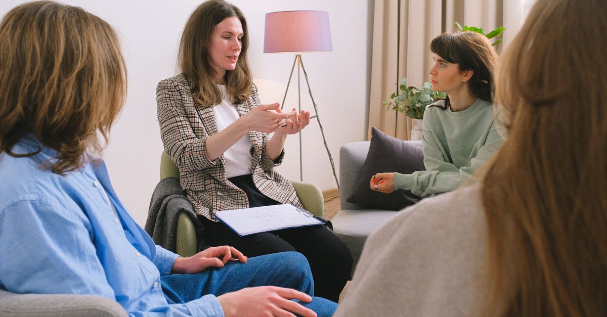 Young female therapist sitting on chair and discussing problem with patients during psychotherapy session in group
