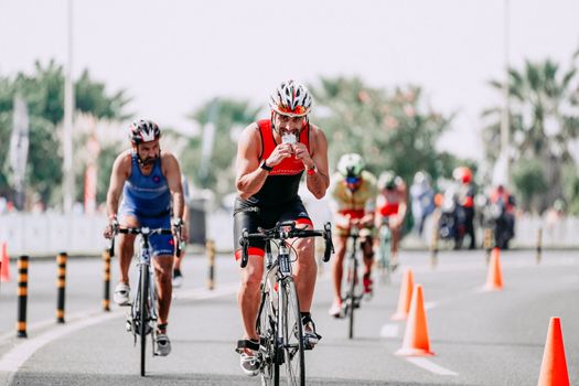 Fit male bicyclist drinking beverage while riding bicycle behind unrecognizable rivals during race on roadway in city