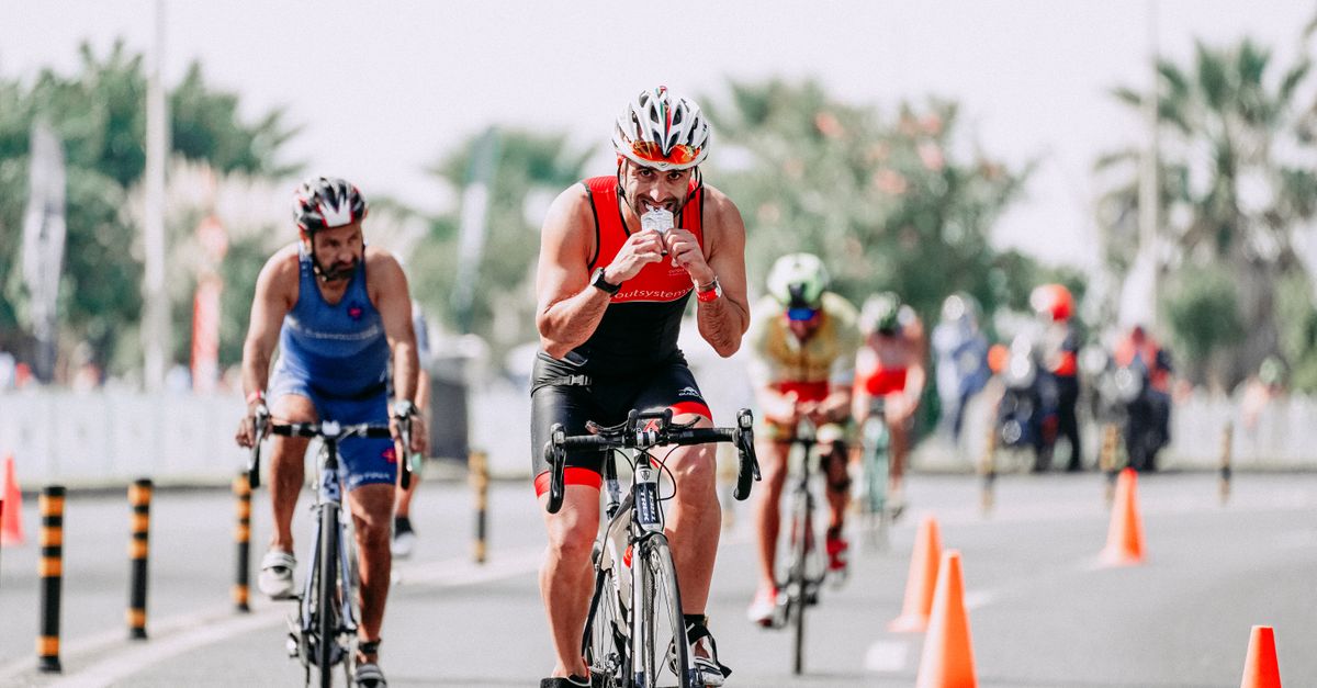 Fit male bicyclist drinking beverage while riding bicycle behind unrecognizable rivals during race on roadway in city