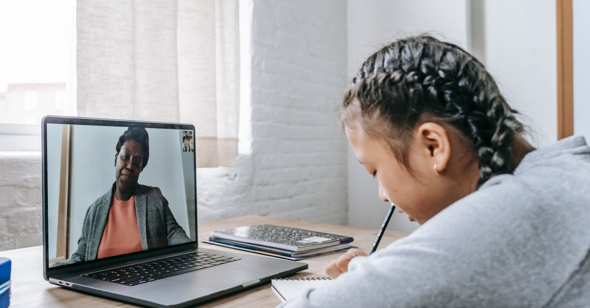 Side view of focused Asian schoolgirl sitting at wooden table and writing notes during remote lesson on laptop with African American teacher