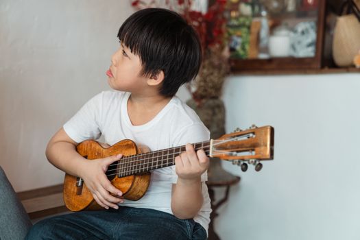 Melancholic ethnic boy in casual wear playing classic musical instrument while sitting near kitchen and looking away at home on blurred background