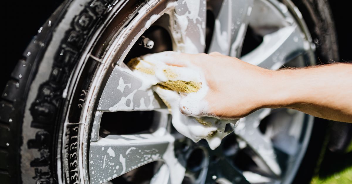 A close-up image of a hand scrubbing a car rim and tire with suds, emphasizing car wash detail.